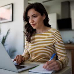 Woman looking at her laptop, researching ENTs and taking notes in a notebook.