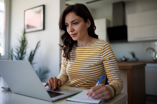 Woman looking at her laptop, researching ENTs and taking notes in a notebook.