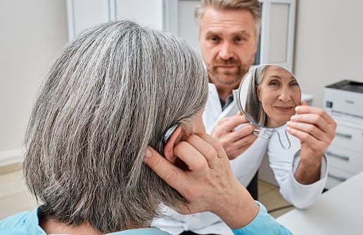 Audiology patient tries on her new hearing aids at the audiologist's office. Her doctor holds up a mirror so she can see her new hearing aid.