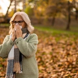 Mature woman sneezing and blowing nose with napkin while on a walk in nature during autumn.
